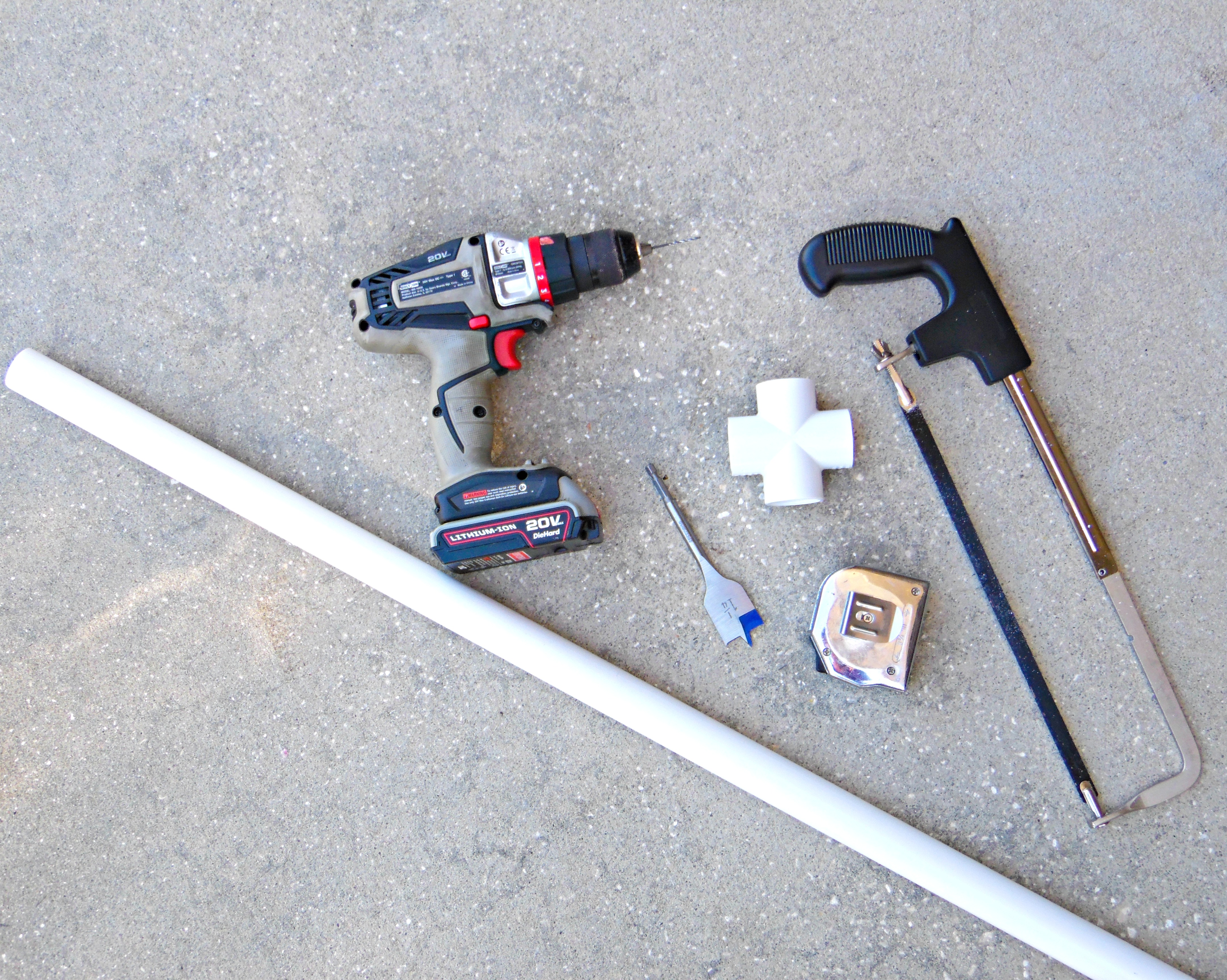 A PVC pipe, drill, and handsaw sit on the concrete outside. These are some of the tools you need to gather to make a free-standing flower stand for your giant paper flower stems. 