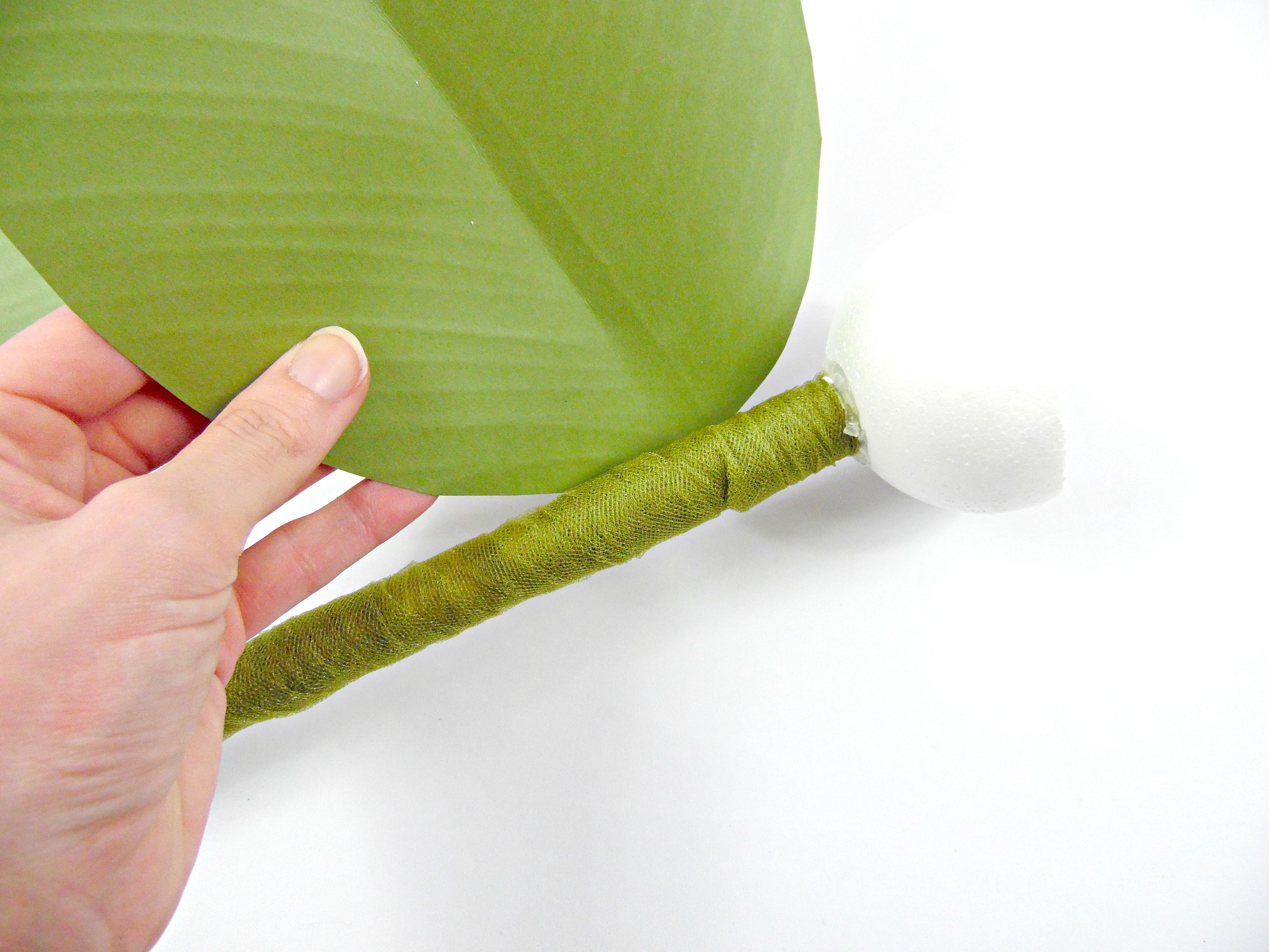 This giant paper flower stem is ready to have the leaves attached. Here, we are attaching one giant paper flower leaf to a tulle-covered stem, just below the foam ball, which will be the head of the flower.