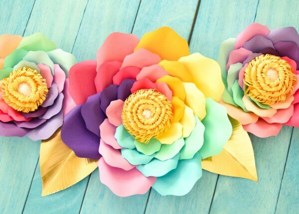 Three finished large rainbow paper flowers on a sea-green wooden table. Petals in pinks, reds, yellows, greens, and purples swirl around the flower to the center yellow fringe. 