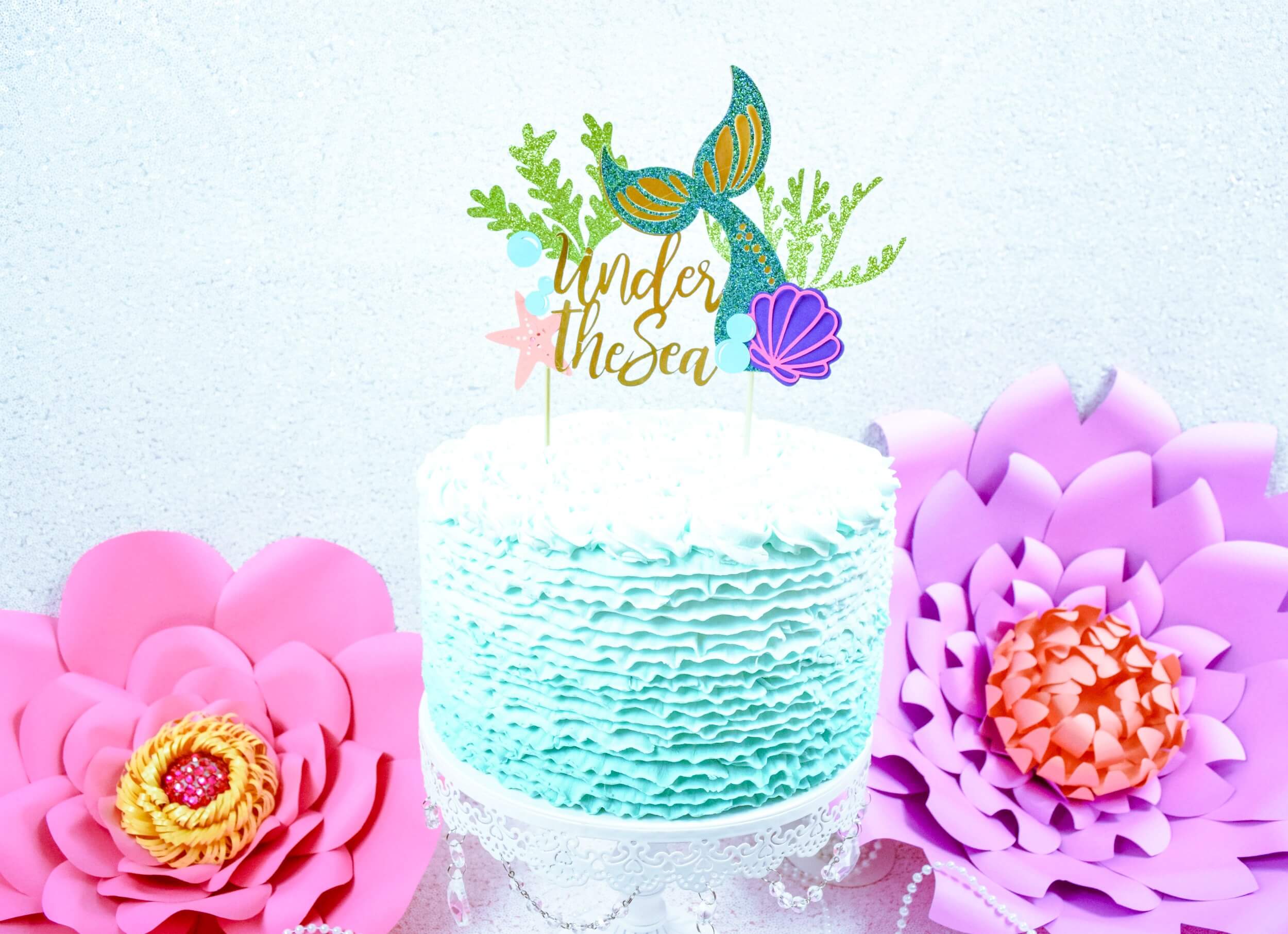 A small round frosted birthday cake sits on a cake stand between two large pink and purple paper flowers. The cake is frosted with a light ombre-blue frosting and topped with a DIY mermaid-themed cake topper that says “under the sea” in gold letters.