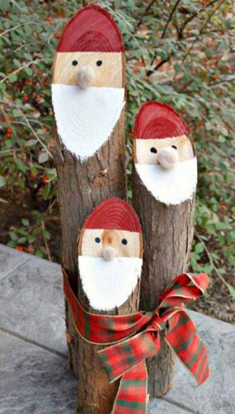 Three wooden stumps are tied together with a plaid ribbon. Each log is cut on an angle at three different lengths, and the end of each log is painted to look like a Santa Clause face.