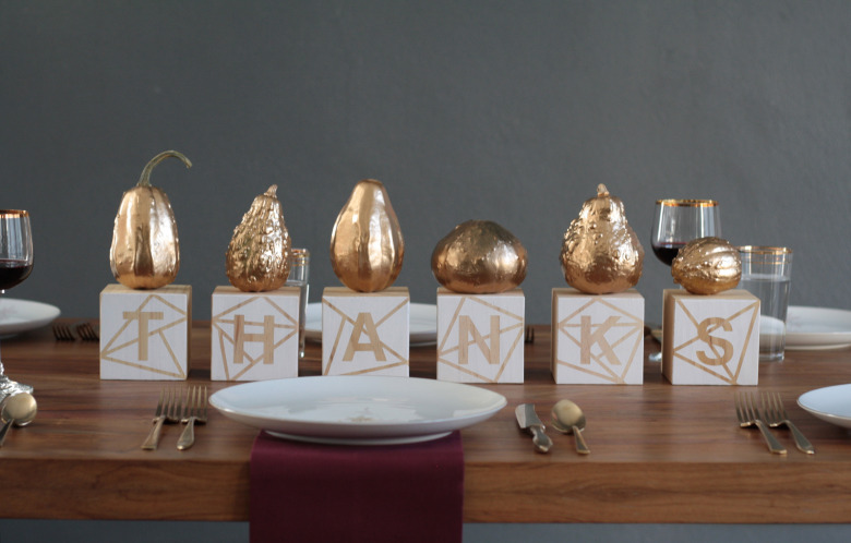 A Thanksgiving themed centerpieces is arranged on a set dining table. Six wood blocks are painted white with a gold geometric pattern, and spell out the word "thanks". Each block has a gold-painted gourd on top.