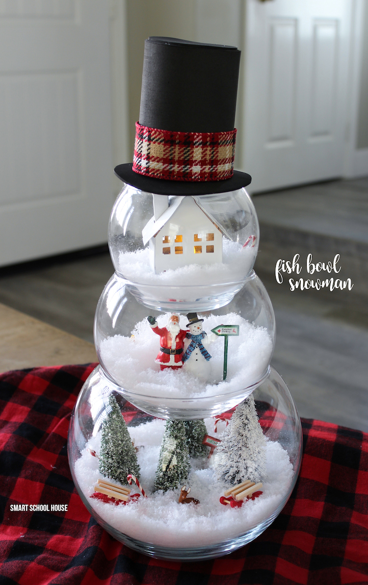 A Christmas snowman is created from three stacked glass fishbowls. Each bowl has a different Christmas scene; the largest on the bottom has a forrest of snow-covered trees, the middle has Santa and a snowman figurine posed together, and the smallest top bowl has a small house. 