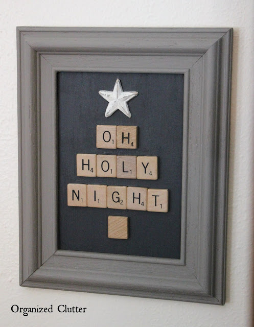 A small "oh holy night" framed sign hangs on a white wall. The sign is made with Scrabble pieces arranged in the shape of a Christmas tree. a single small wooden start tops the tree.