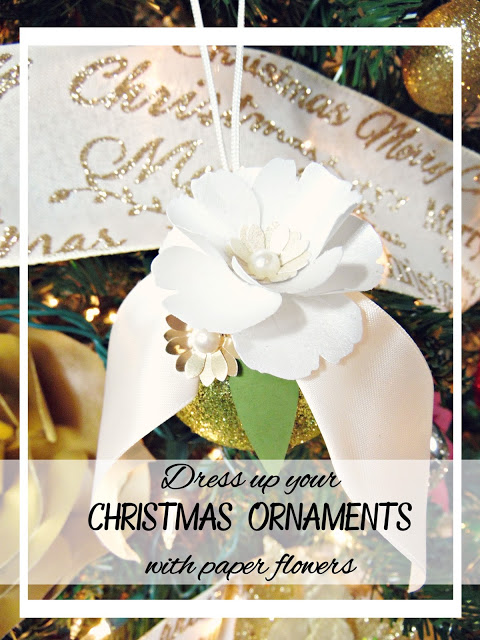 A gold globe ornament topped with a white ribbon and white paper flower hangs from a Christmas tree with lights.