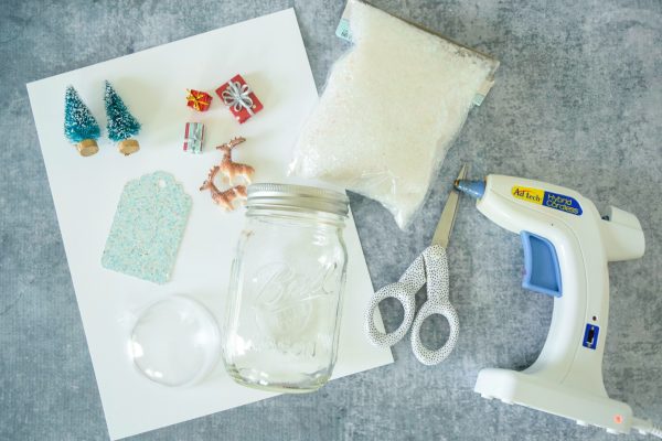 A helpful overhead view of supplies needed to make mason jar Christmas snowglobes. Featured are scissors, a glue gun, mini holiday props, a mason jar, and more. 
