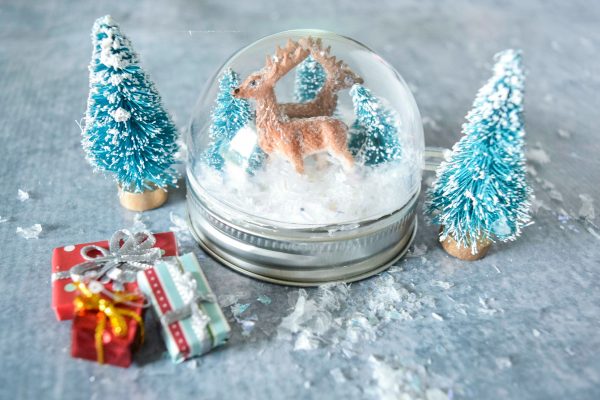 A closeup of a snowglobe on top of a mason jar lid featuring a deer,  Christmas trees, and tiny presents.