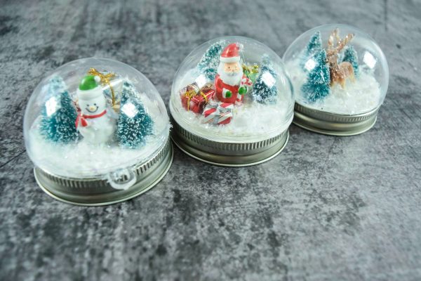 Three different Christmas scenes made out of holiday mini figurines are captured in snowglobes glued to mason jar lids. 