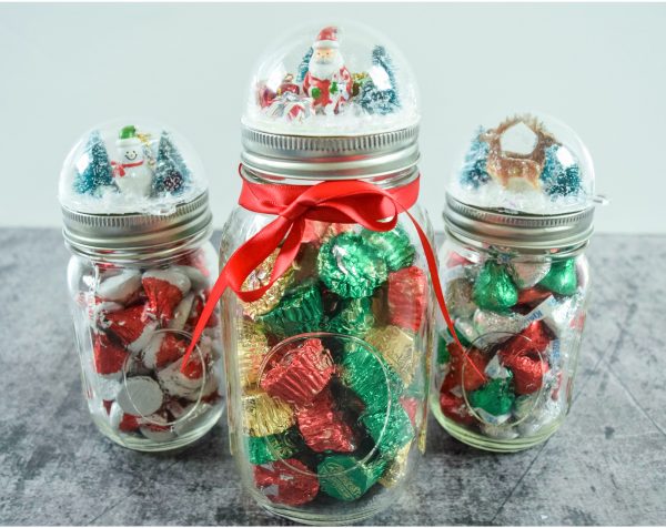 On a marble table sit three mason jars full of Christmas candy. On top of each mason jar is a DIY snowglobe with holiday scenes. 