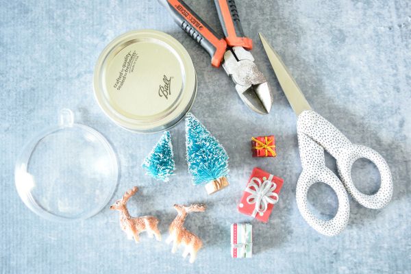 An acrylic dome, a mason jar lid, tiny Christmas scene decor, and scissors are some of the supplies needed to make DIY mason jar snowglobe crafts. 
