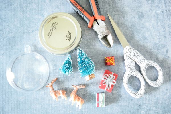 An acrylic dome, a mason jar lid, tiny Christmas scene decor, and scissors are some of the supplies needed to make DIY mason jar snowglobe crafts. 