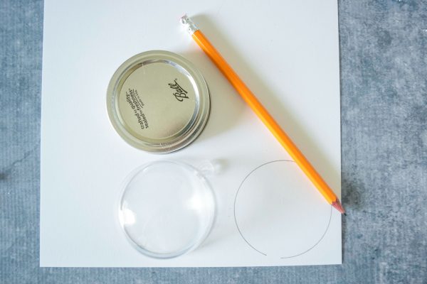 A piece of white cardstock lays on a gray tabletop. On the paper is a pencil, a mason jar lid, and half of a clear fillable ornament. 