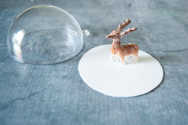 A half dome of clear acrylic is next to a cardstock circle with a miniature deer figurine. These are the first couple of steps in making a DIY snowglobe mason jar craft.