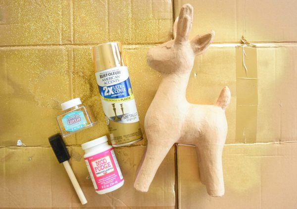 A paper mache deer laying on a cardboard piece with gold spray paint and other supplies. 