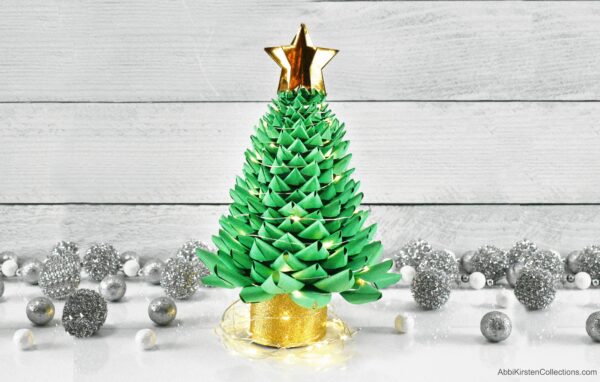 A small green paper Christmas tree had a gold base and a gold star. Fairy lights are wrapped around the base and the tree. Silver and white round ornaments decorate the white table. Download the FREE SVG and PDF printable templates.