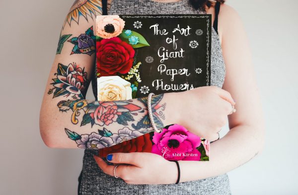 A professional crafter with colorful flower tattoos on her arm cradles Abbi Kirsten's book "The Art of Giant Paper Flowers." 