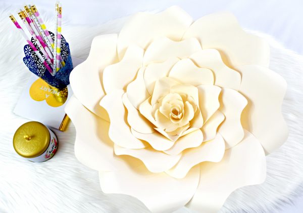 A finished Bella Rose flower, made using a Cricut machine and paper. Pencils and other craft supplies show the large size of the white craft flower. 