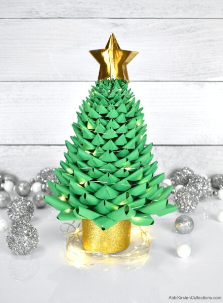 A finished green paper 3D Christmas tree. Silver and white round decorations adorn the white tabletop and white wooden wall. The holiday tree has fairy lights, a gold circular base and a 6 layer 3D paper star topper. 