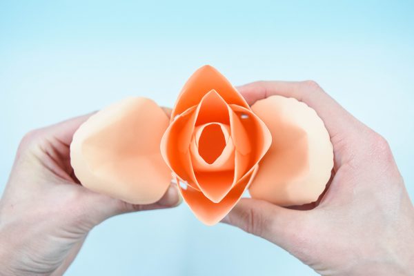A woman's hands hold a large paper rose center as she presses new petals onto the established bud. The DIY paper rose is a peach serena rose.