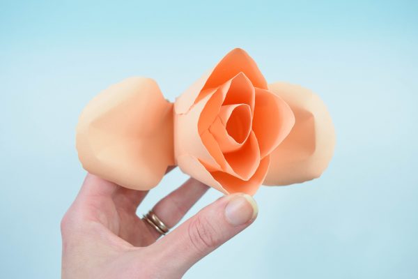 Abbi Kirsten's hand holds the bud of a paper rose with shaped and curled petals attached and ready to be added to the bud. 
