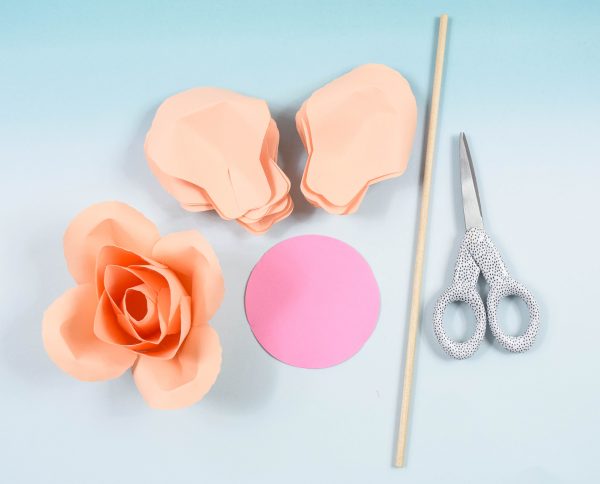 An overhead view of the supplies needed to make DIY paper roses. The serena rose requires petals cut from cardstock, a thin dowel, scissors, a small paper circle, and the start of the rosebud center. 