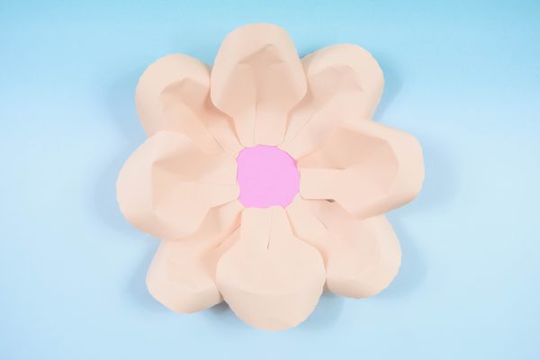 On a baby blue background, a pink paper circle has eight large paper petals glued and overlapped around its edges. 