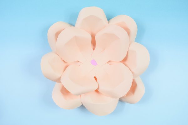 Two layers of a peach DIY paper rose serena flower is seen from above, sitting on a light blue tabletop. 