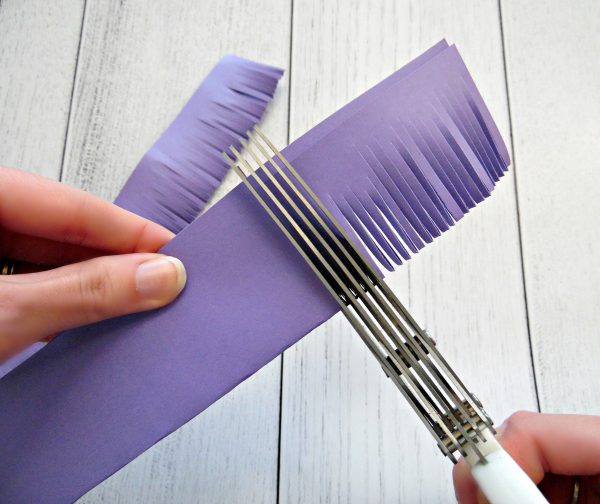 A closeup view of Abbi Kirsten's hands as she uses shredding scissors to create fringe on the edges of folded purple paper. 