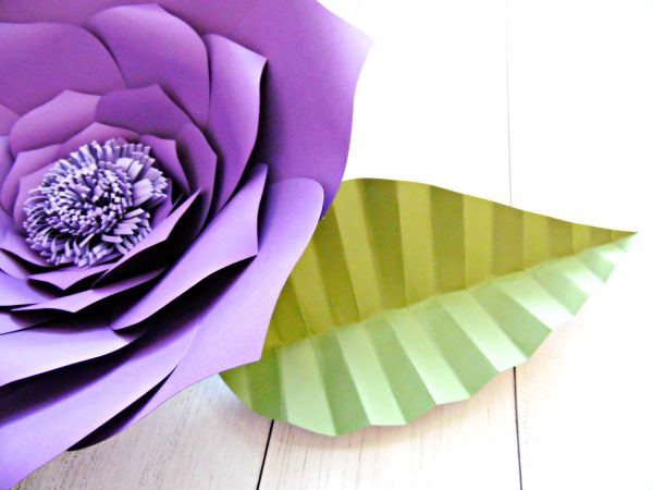 The open green paper leaf with accordion folds accents the right side of a giant purple paper flower on a white table top. Get the template from AbbiKirstenCollections.