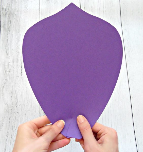 A closeup of Abbi Kirsten's hands holding two sides of a paper petal together to form a curl at the base. The giant purple paper petal is part of the assembly of a giant paper flower cut by a Cricut cutting machine. 