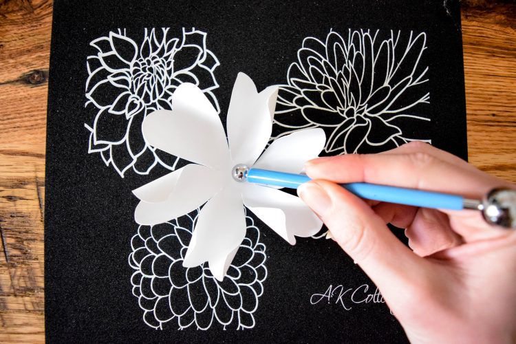 Image shows Abbi using rolling tools to roll and curl a white paper blower on a black crafting mat.