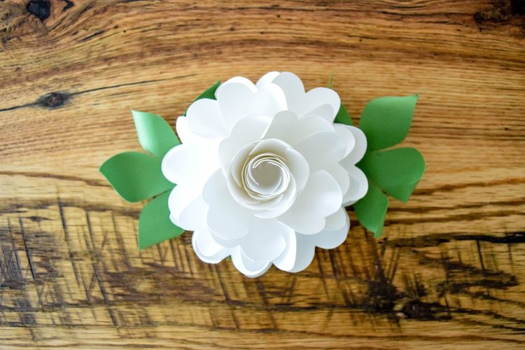 A completed white paper pomander flower ball with green leaves, sitting on a wooden surface.