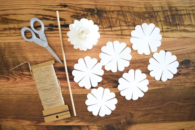 Six pomander paper flower templates cut out, and one completed pomander flower, a wooden dowel, scissors, and twine set out on a wooden table.