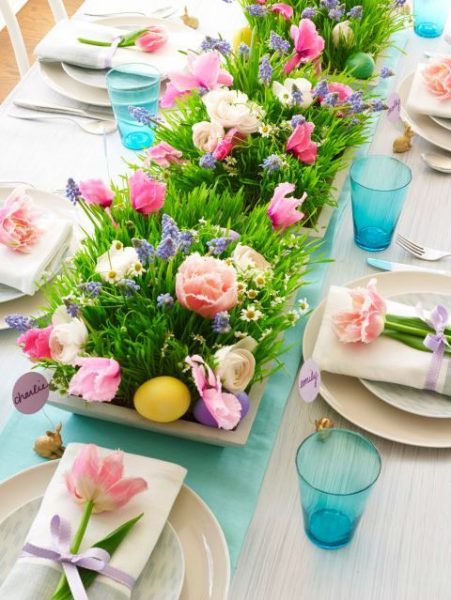 A table centerpiece with grass, pastel flowers and eggs. There are place settings around the table. 