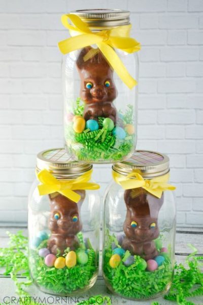 Three clear mason jars filled with chocolate Easter bunnies, green paper grass and pastel candies. The jars are tied with a yellow ribbon. 