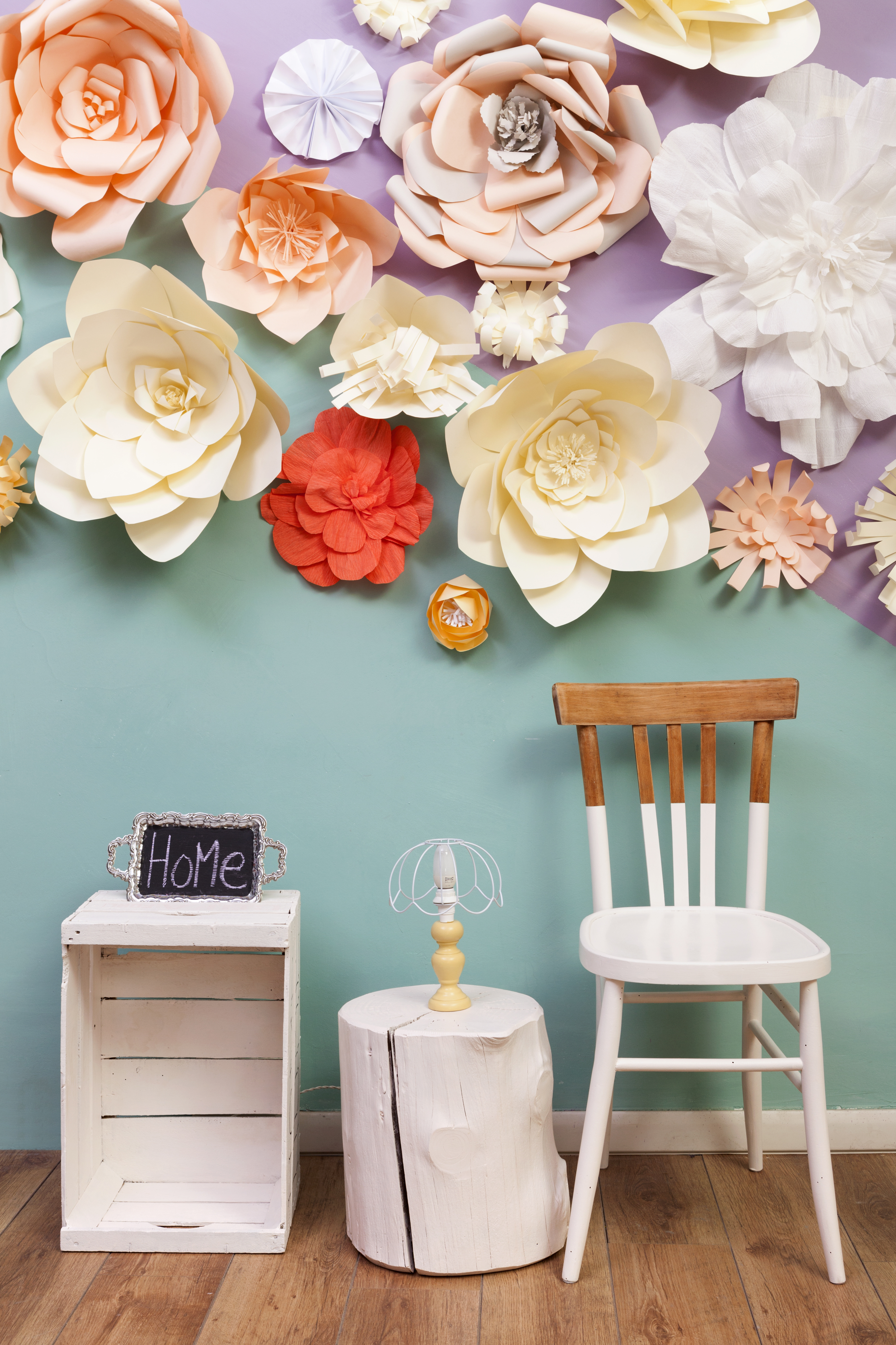 Peach, tan, orange, and white giant paper flowers hang on a green and purple wall, over a wooden chair and side table set up.