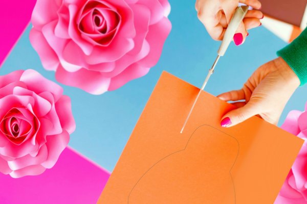 A woman's hands use scissors to cut out a paper flower petal from orange cardstock. Pink paper flowers adorn the workspace. 