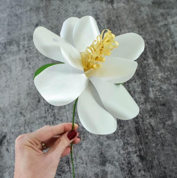 A woman's manicured hand holds a large white Magnolia Paper Flower, with ten white flower petals, a yellow center, and green wire stem.