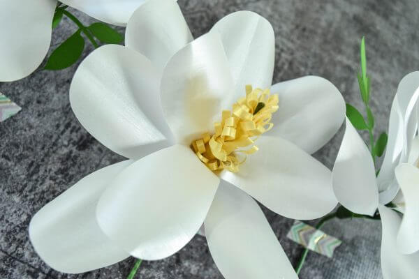A completed Southern Magnolia Paper Flower made with white petals and a yellow center, laying flat on a black background.