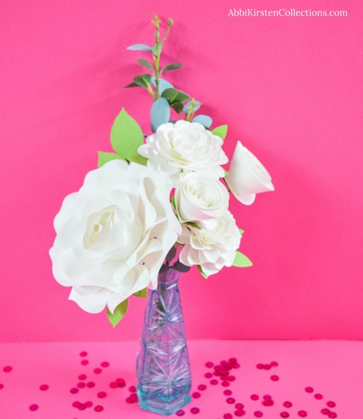 White paper flowers in a clear blue glass vase against a bright pink background with red sequin confetti. Learn what supplies you need to make paper flowers. 