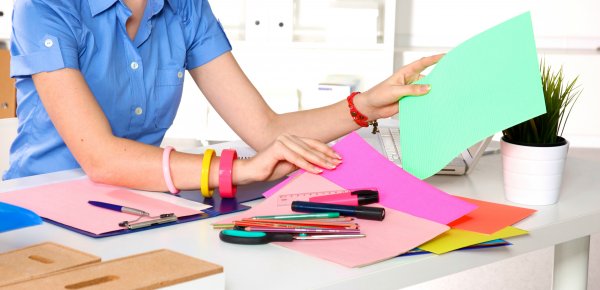 A woman sits at a desk, sorting through paper and markers for crafting. If you love paper flowers, find out what supplies you need from Abbi Kirsten. 