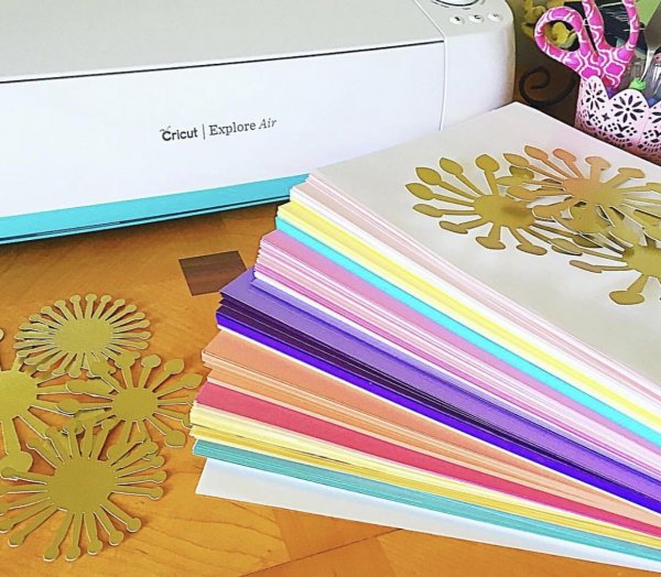 A close view of a Cricut Explore Air, a fanned-out stack of cardstock, and some gold cutouts in the shape of asterisks and stars. 