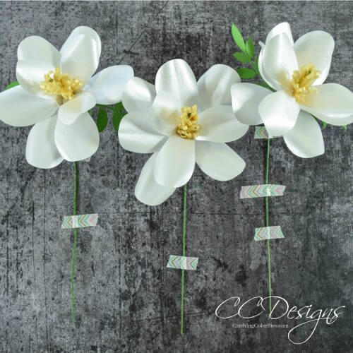 Three white Magnolia Paper Flowers with yellow centers and green stems, laying against a black background.