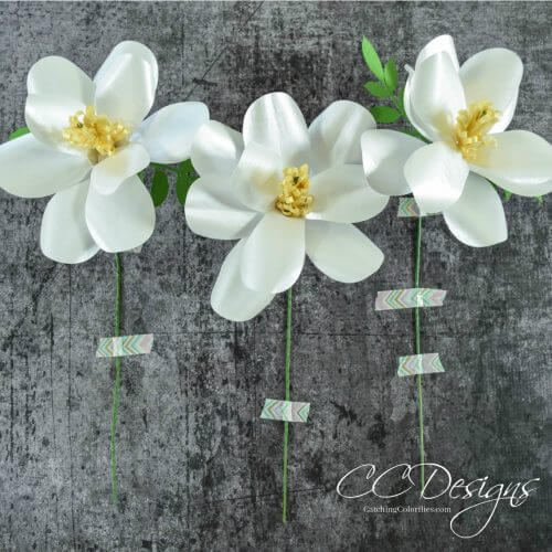 Three delicate paper southern magnolia flowers are taped to a dark gray table by their thin stems. The corner text says “CC Designs.”