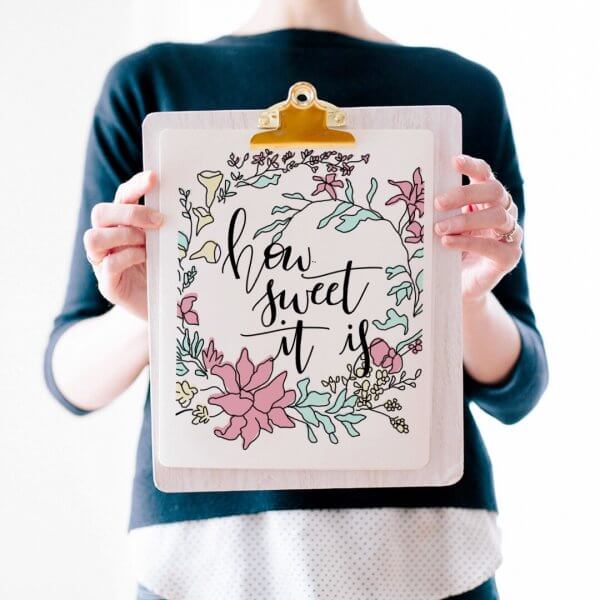 Abbi holds up a free Spring Printable that reads "how sweet it is" in black text surrounded by a ring of pastel spring flowers.