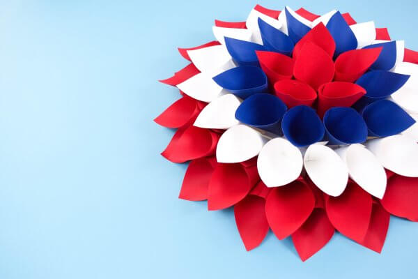 A patriotic paper dahlia wreath made with cones of red, white, and blue paper layered together, sitting on a light blue background.