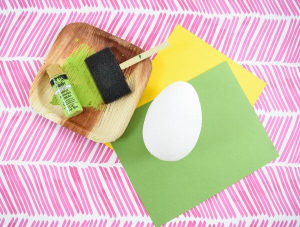 An overhead view of a faux wooden square paper plate with a foam paint brush and green paint in it. Next to the paint is a foam egg on top of green and yellow cardstock. 