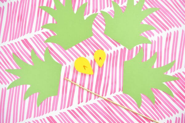 Four green paper pineapple stems, two yellow paper pineapple scales and a bamboo skewer lay on a background of fun pink slashes. 