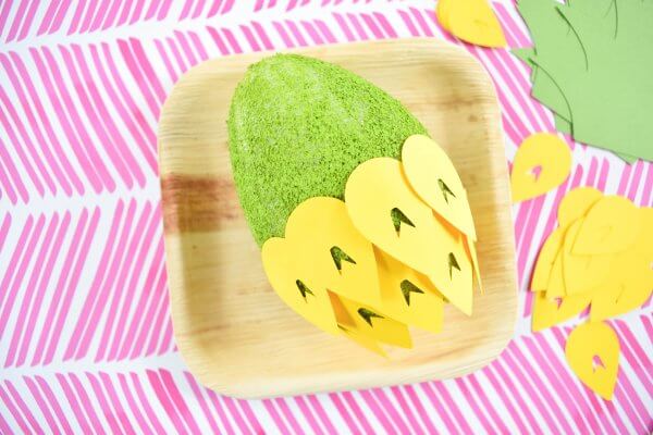 In a clean fake wooden square paper plate sits a neon-green painted foam egg half covered with yellow paper pineapple scales. Additional scales and paper stem parts sit off to the side. 