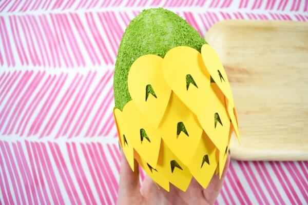 A green foam egg is almost completely covered with yellow paper pineapple scales. The background is pink slashes and a fake wooden square paper plate. 